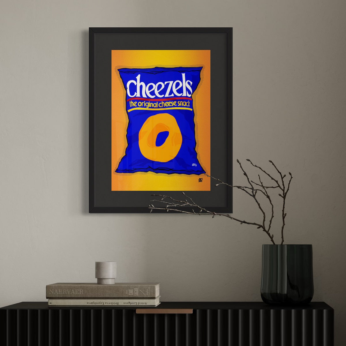 Framed artwork of a blue Cheezels bag on an orange background, displayed on a wall above a black shelf.