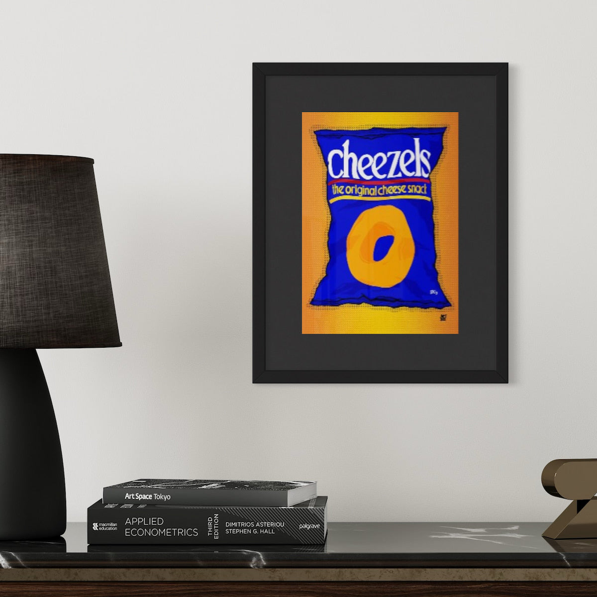 Framed artwork of a Cheezels bag on a shelf with books and a lamp.