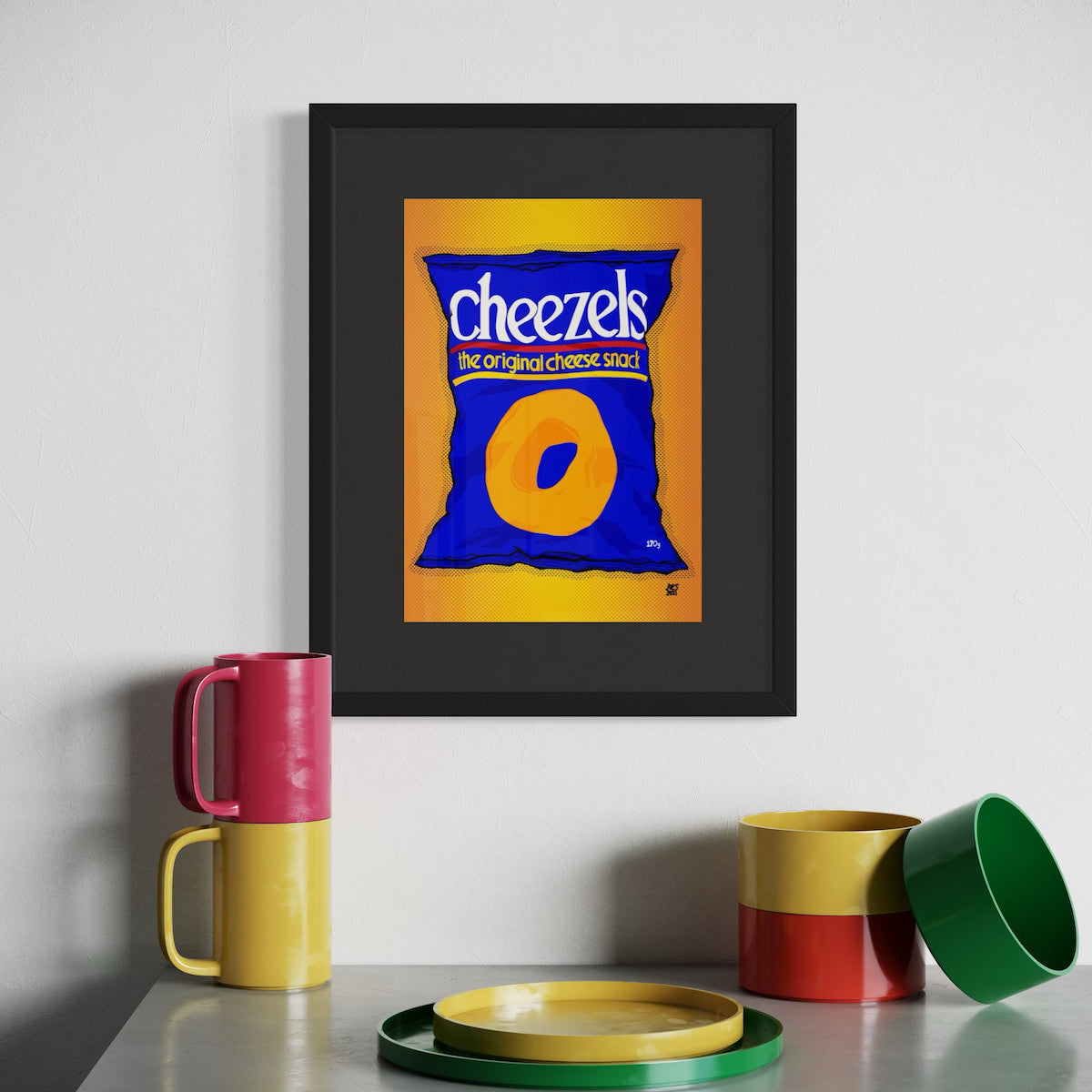 Framed artwork of a Cheezels bag on a wall with colorful mugs and a tray below.