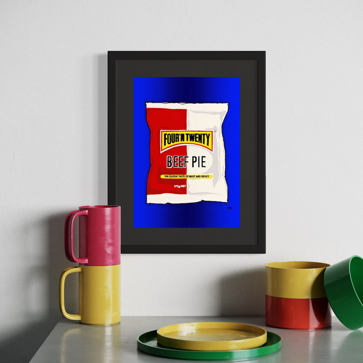 Framed artwork of a beef pie package with colorful mugs and plates on a table.