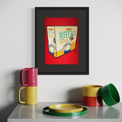 Framed artwork of a Weet-Bix cereal box on a wall above colorful mugs and a plate.