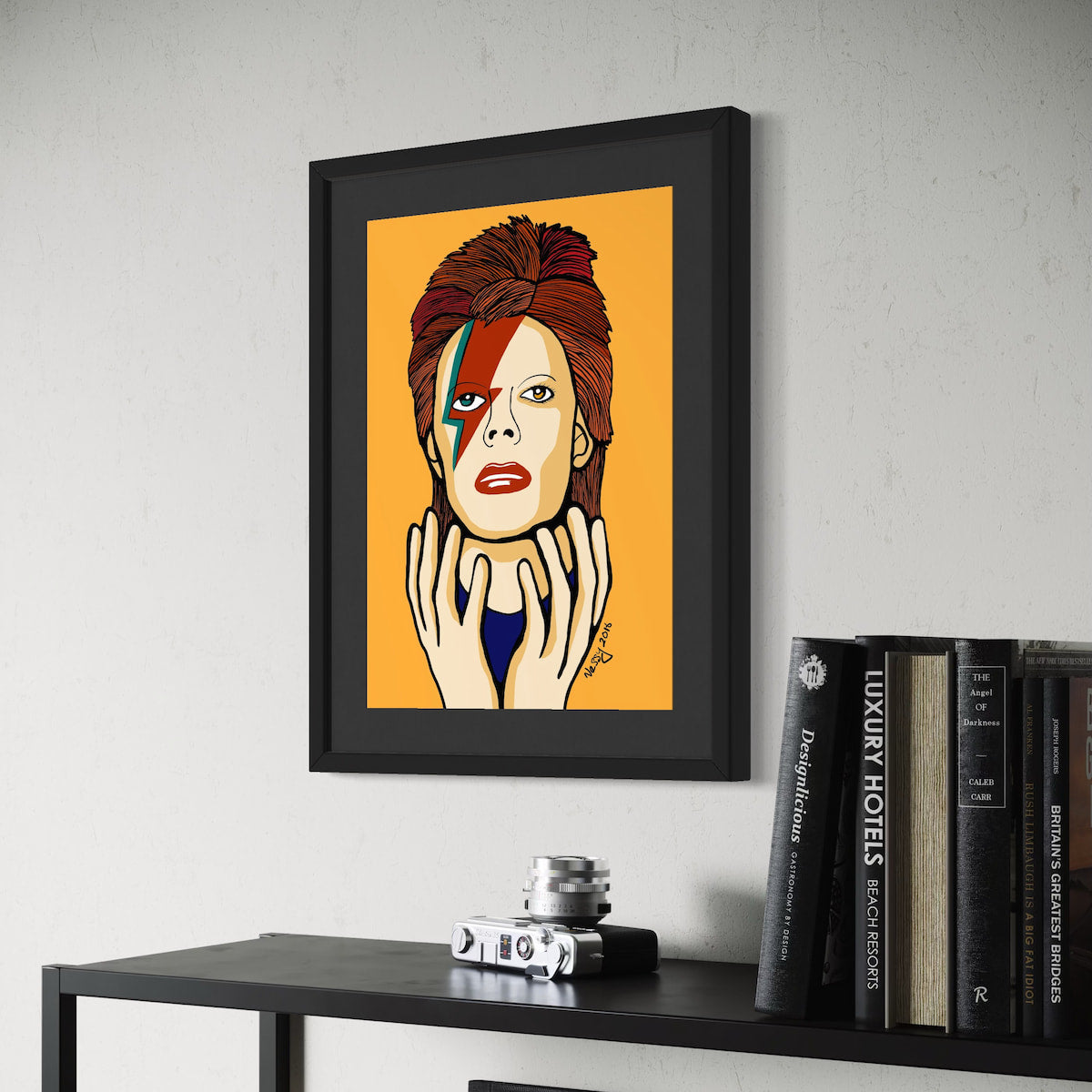 Framed artwork of David Bowie with a colorful design on an orange background, displayed on a wall above a shelf with books and a camera.